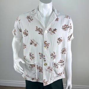 May include: White short-sleeved shirt with a brown and tan tropical print. The shirt has two front pockets and a button-up closure. The collar and pocket edges have a textured trim.