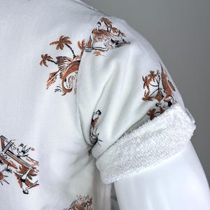 May include: White short-sleeved shirt with a brown and black tropical print. The shirt features a rolled-up sleeve with a white terry cloth cuff. The print includes palm trees, boats, and figures.