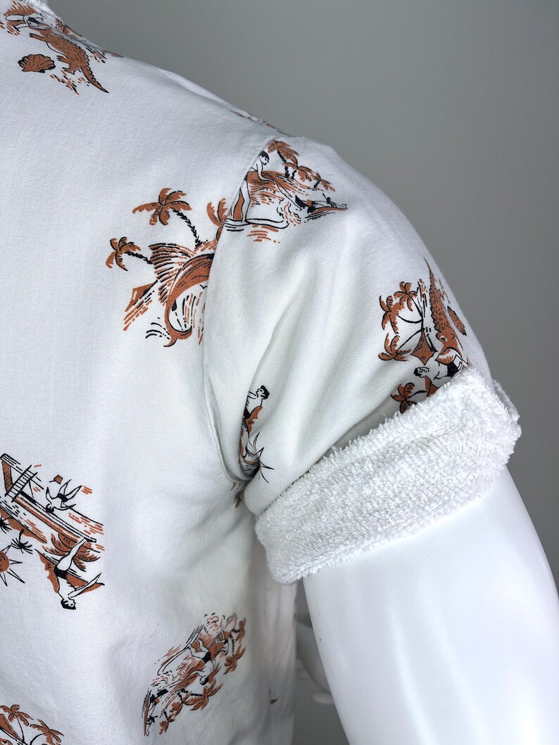 May include: White short-sleeved shirt with a brown and black tropical print. The shirt features a rolled-up sleeve with a white terry cloth cuff. The print includes palm trees, boats, and figures.
