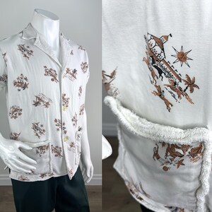 May include: White short-sleeve shirt with a camp collar and a brown and tan print. The shirt has two front pockets and a button-up closure. The print features various figures and shapes.