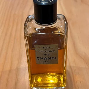 Puede incluir: Una botella rectangular de vidrio de Chanel No. 5 Eau de Cologne. La botella contiene un l&iacute;quido dorado y tiene una tapa negra con un logotipo dorado. La etiqueta dice "EAU DE COLOGNE N&deg;5 CHANEL PARIS."