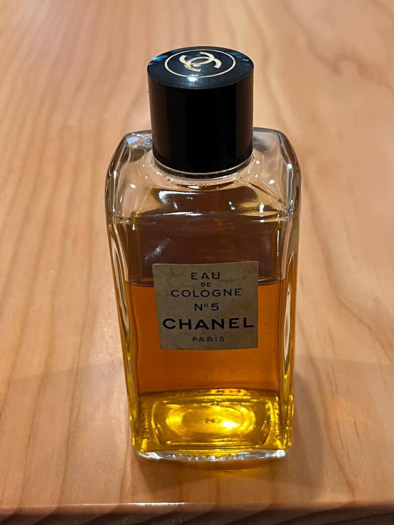 Puede incluir: Una botella rectangular de vidrio de Chanel No. 5 Eau de Cologne. La botella contiene un l&iacute;quido dorado y tiene una tapa negra con un logotipo dorado. La etiqueta dice "EAU DE COLOGNE N&deg;5 CHANEL PARIS."