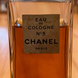 Puede incluir: Una botella rectangular de vidrio de Chanel No. 5 Eau de Cologne. La botella contiene un l&iacute;quido &aacute;mbar oscuro. Una etiqueta rectangular en la parte delantera dice "EAU DE COLOGNE N&deg;5 CHANEL PARIS."