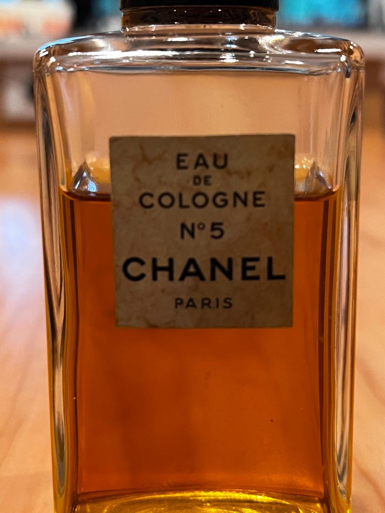 Puede incluir: Una botella rectangular de vidrio de Chanel No. 5 Eau de Cologne. La botella contiene un l&iacute;quido &aacute;mbar oscuro. Una etiqueta rectangular en la parte delantera dice "EAU DE COLOGNE N&deg;5 CHANEL PARIS."