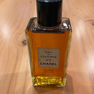 K&ouml;nnte beinhalten: Eine Vintage-Flasche Chanel No. 5 Eau de Cologne. Die rechteckige Glasflasche enth&auml;lt eine goldfarbene Fl&uuml;ssigkeit und hat einen schwarzen Verschluss mit einem Logo. Auf dem Etikett steht "Eau de Cologne N&deg;5 Chanel".