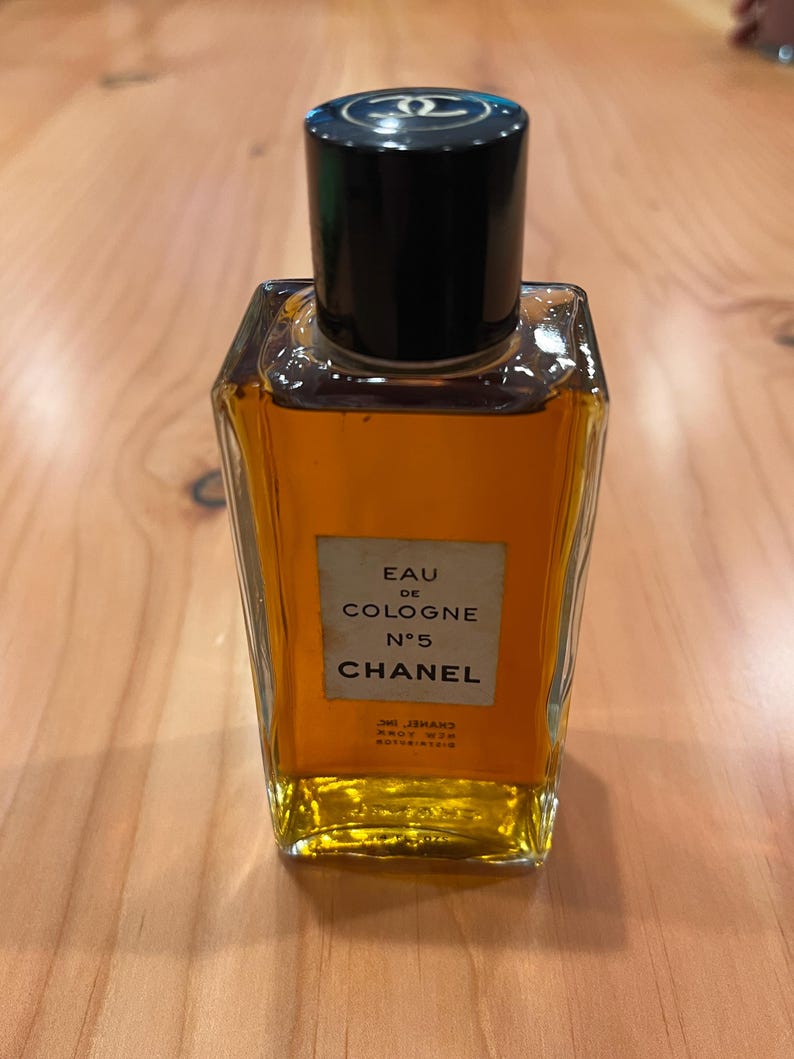 K&ouml;nnte beinhalten: Eine Vintage-Flasche Chanel No. 5 Eau de Cologne. Die rechteckige Glasflasche enth&auml;lt eine goldfarbene Fl&uuml;ssigkeit und hat einen schwarzen Verschluss mit einem Logo. Auf dem Etikett steht "Eau de Cologne N&deg;5 Chanel".