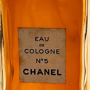 K&ouml;nnte beinhalten: Nahaufnahme einer Vintage-Flasche Chanel No. 5 Eau de Cologne. Die bernsteinfarbene Fl&uuml;ssigkeit ist durch das klare Glas sichtbar. Ein rechteckiges Etikett zeigt den Produktnamen in schwarzer Schrift: "EAU DE COLOGNE N&deg;5 CHANEL."