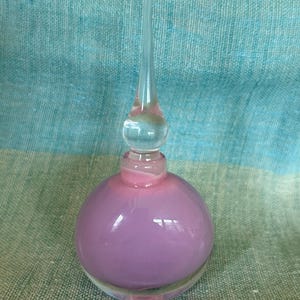 May include: A round, lavender glass perfume bottle with a clear, elongated stopper. The bottle has a pink ombre effect, transitioning from lavender to pink. The base is clear glass, and the stopper is a long, clear, pointed design.