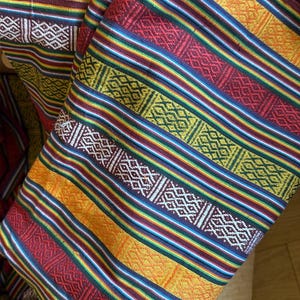 May include: Close-up of a folded textile with a vibrant striped pattern. The fabric features alternating bands of red, yellow, blue, and green, with intricate geometric designs in white and burgundy. The textile is likely used for clothing or home decor.