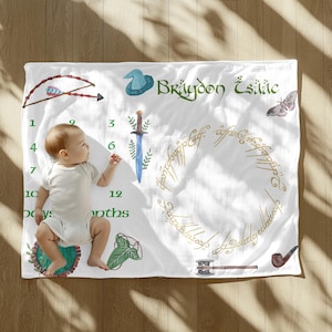 Puede incluir: Manta blanca para bebé con detalles en verde y dorado. Presenta ilustraciones de un arco y una flecha, un sombrero de mago, una espada y un anillo con texto. Incluye el nombre "Brýdon Īsaac" y un bebé.