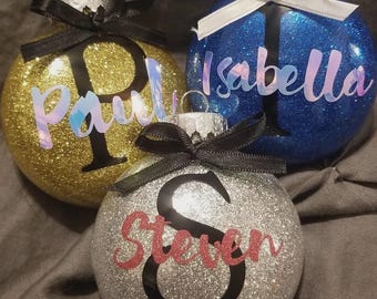 May include: Three glitter-covered Christmas ornaments. One is gold with "Paul" in iridescent lettering, another is blue with "Isabella" in iridescent lettering, and the last is silver with "Steven" in red lettering. Each has a black or white bow.