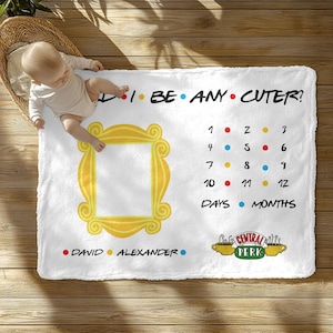 May include: White baby milestone blanket with the text "I BE ANY CUTER?" and a yellow frame. Includes a calendar for tracking baby's age in days and months. Features the names "DAVID" and "ALEXANDER" and the "Central Perk" logo.