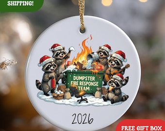 May include: A round white ceramic ornament featuring a cartoon illustration of five raccoons wearing Santa hats around a green dumpster fire. The dumpster is labeled DUMPSTER FIRE RESPONSE TEAM. The year 2026 is printed below the illustration.