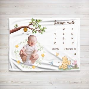 May include: White baby milestone blanket featuring a smiling baby surrounded by a floral wreath. The blanket includes a tree branch with a beehive, Winnie the Pooh and Piglet, and a space to track baby's growth in days and months.