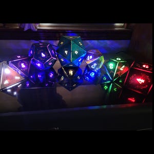 May include: A set of seven polyhedral dice featuring intricate floral border engravings around the numbers. Each die is illuminated from within, emitting vibrant blue, green, and red light that highlights the etched details on the dark surfaces.