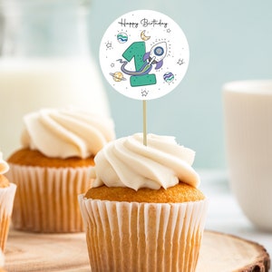 Op de afbeelding: Een cupcake met witte frosting bedekt met een ronde, witte papieren cupcake topper met de tekst "Happy Birthday" en een groen nummer één met een raket en sterren.