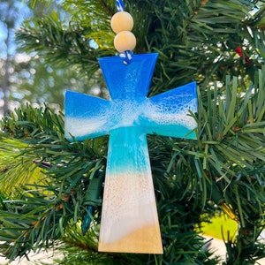 Op de afbeelding: Een blauwe en witte hars kruis ornament met een strand sc&egrave;ne, hangend aan een kerstboom. Het kruis is versierd met een wit en blauw golf ontwerp en een bruine zand bodem.