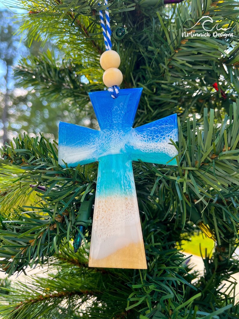 Op de afbeelding: Een blauwe en witte hars kruis ornament met een strand sc&egrave;ne, hangend aan een kerstboom. Het kruis is versierd met een wit en blauw golf ontwerp en een bruine zand bodem.