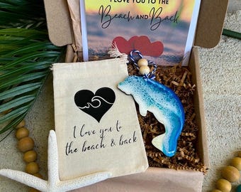 Op de afbeelding: Een geschenkdoos met een klein beige trekkoordzakje met een zwart hart- en golfmotief en de tekst I love you to the beach & back, een blauw-witte dolfijnornament van hars en een kaart met de tekst I LOVE YOU TO THE BEACH AND BACK.