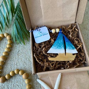Pode incluir: Um ornamento de barco &agrave; vela de madeira azul e branco com um design de onda branca. O ornamento est&aacute; em uma caixa de papel&atilde;o marrom com material de embalagem de papel marrom. A caixa tem uma etiqueta branca com o texto "Minnick Designs" nela.