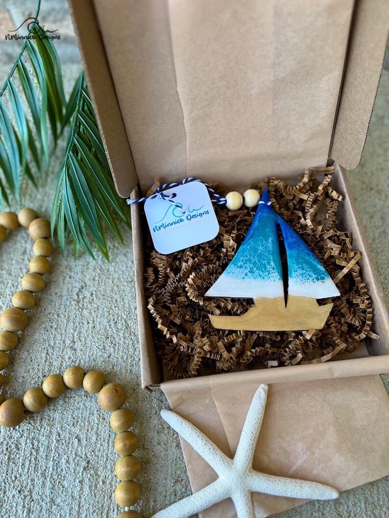 Pode incluir: Um ornamento de barco &agrave; vela de madeira azul e branco com um design de onda branca. O ornamento est&aacute; em uma caixa de papel&atilde;o marrom com material de embalagem de papel marrom. A caixa tem uma etiqueta branca com o texto "Minnick Designs" nela.