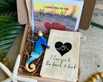 Op de afbeelding: Een kartonnen geschenkdoos met een blauw en wit zeepaardje ornament van hars met een blauw en wit koord, een wit stoffen zakje met een zwart hart en golfafbeelding, en een kaart met de tekst I LOVE YOU TO THE Beach AND Back.