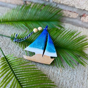 Pode incluir: Um ornamento de barco &agrave; vela de madeira pintado em tons de azul e branco com um efeito de branqueamento. O ornamento est&aacute; pendurado em um cord&atilde;o azul e branco com contas de madeira.