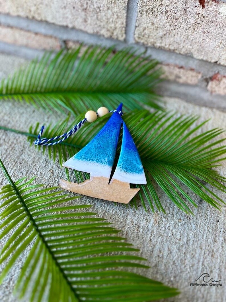 Pode incluir: Um ornamento de barco &agrave; vela de madeira pintado em tons de azul e branco com um efeito de branqueamento. O ornamento est&aacute; pendurado em um cord&atilde;o azul e branco com contas de madeira.