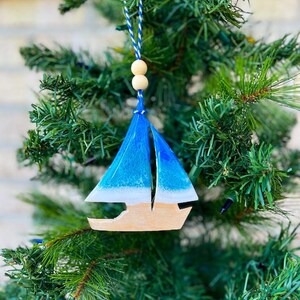 Pode incluir: Um enfeite de Natal de veleiro azul e branco com base de madeira. O enfeite est&aacute; pendurado em uma &aacute;rvore de Natal artificial verde.
