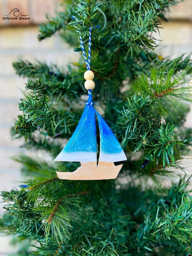 Pode incluir: Um enfeite de Natal de veleiro azul e branco com base de madeira. O enfeite est&aacute; pendurado em uma &aacute;rvore de Natal artificial verde.
