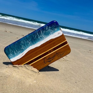 May include: A wooden cutting board with a blue and white resin wave design on one end. The board has a shark graphic and is resting on a sandy beach. The board is made of wood with a dark brown and light brown color scheme.