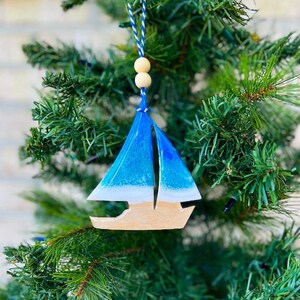 A christmas ornament shaped like a sailboat adorned with a delicate blue and teal ocean resin wave design and hanging on a christmas tree.