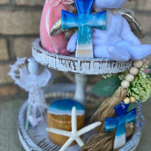 Op de afbeelding: Een wit etag&egrave;re met twee niveaus met een blauw en wit kruis, een witte teddybeer, een roze konijn, een zeester, een houten doos met een blauw en wit deksel en een witte kwast met een blauw en wit kruis.