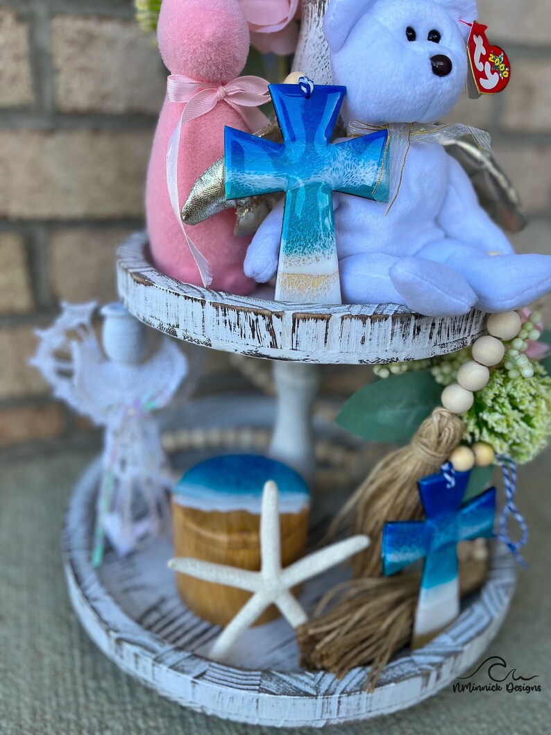 Op de afbeelding: Een wit etag&egrave;re met twee niveaus met een blauw en wit kruis, een witte teddybeer, een roze konijn, een zeester, een houten doos met een blauw en wit deksel en een witte kwast met een blauw en wit kruis.
