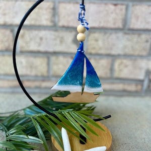 Pode incluir: Um ornamento de veleiro azul e branco com uma base de madeira e um suporte de metal preto. O veleiro est&aacute; pendurado em um cord&atilde;o branco e azul com contas de madeira.