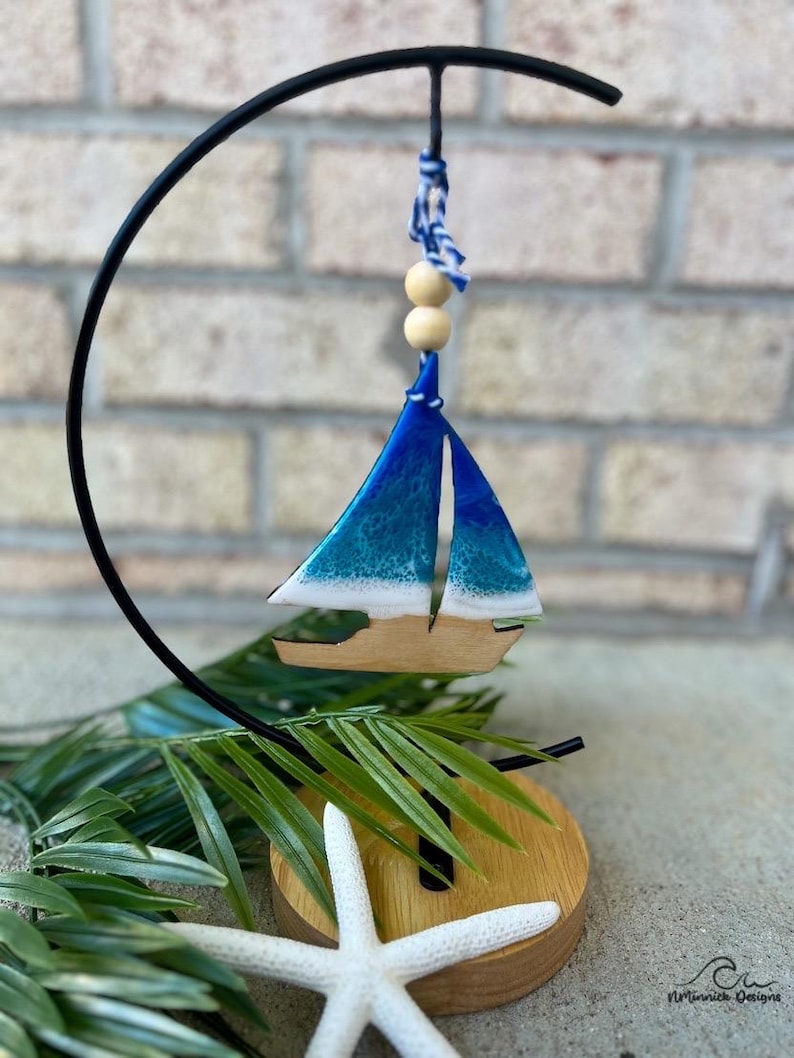 Pode incluir: Um ornamento de veleiro azul e branco com uma base de madeira e um suporte de metal preto. O veleiro est&aacute; pendurado em um cord&atilde;o branco e azul com contas de madeira.