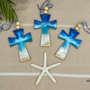 Three cross shaped wooden Christmas ornaments covered in blue ocean resin art, resembling ocean waves.  Finished with baker's twine and two wooden beads.  Laying on a tan beach towel next to a natural starfish and palm leaves.