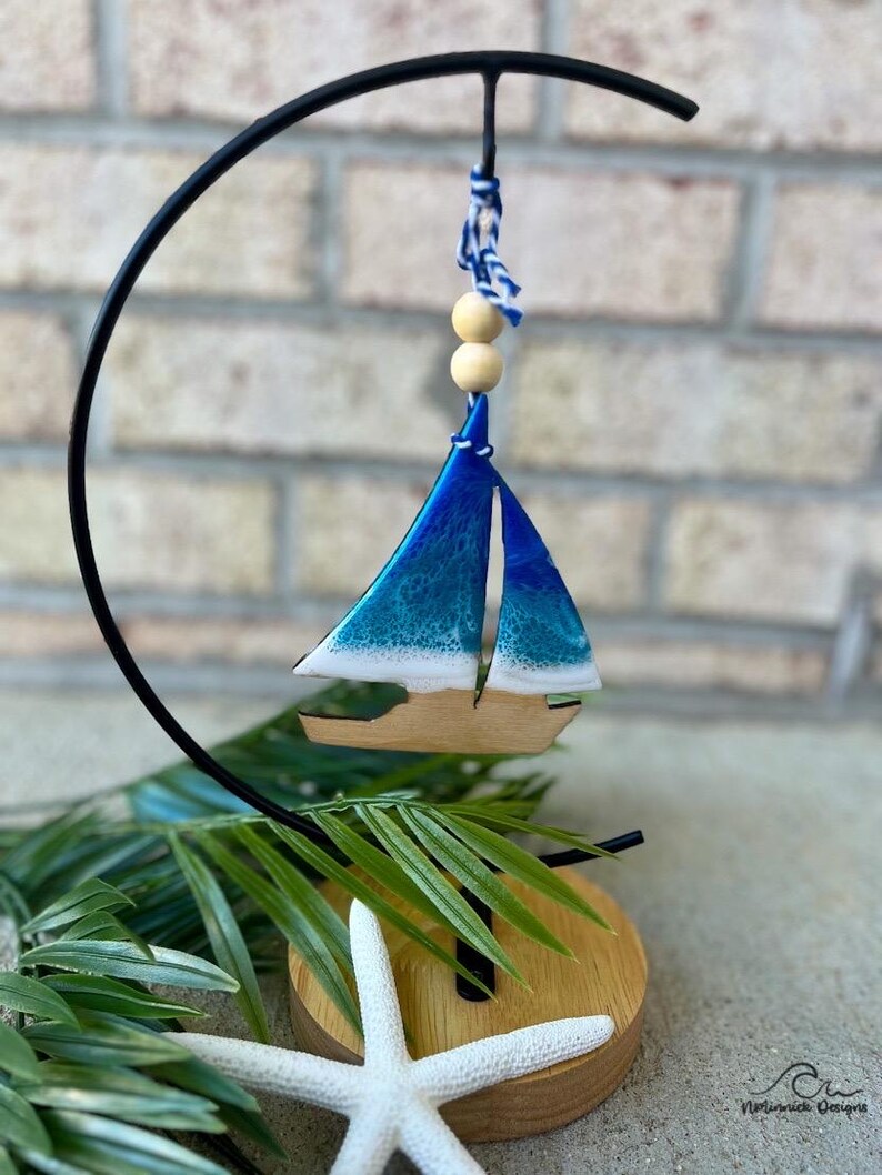 Pode incluir: Um ornamento de veleiro azul e branco com uma base de madeira e uma estrela do mar branca. O ornamento est&aacute; pendurado em um suporte de metal preto.