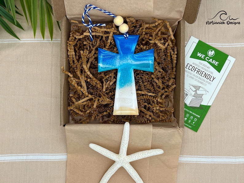 Op de afbeelding: Een blauw en wit kruis met een strandontwerp wordt getoond in een bruine doos met bruine papieren verpakkingschips. Een witte zeester is ook inbegrepen in de doos. De doos heeft een klein kaartje met de tekst "We Care Ecofriendly".