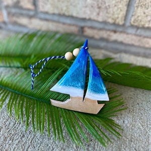 Pode incluir: Um ornamento de barco &agrave; vela de madeira azul e branco com uma corda branca e contas de madeira. O ornamento est&aacute; sobre uma folha de palmeira verde.