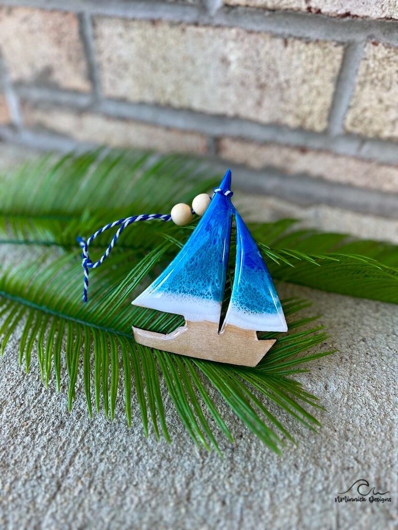 Pode incluir: Um ornamento de barco &agrave; vela de madeira azul e branco com uma corda branca e contas de madeira. O ornamento est&aacute; sobre uma folha de palmeira verde.