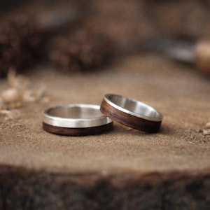 May include: Two wedding rings with a silver band and a dark wood inlay are displayed on a wooden surface. The rings are slightly angled, showcasing the contrast between the silver and the wood. The background is blurred, with wood shavings and a carving tool.