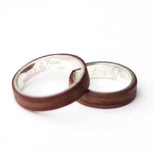 Op de afbeelding: Twee houten ringen met een zilveren band erin. De ringen zijn gegraveerd met de tekst "Emy & Peter".