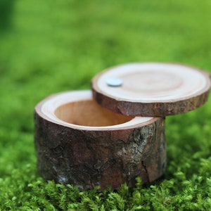 Puede incluir: Una pequeña caja de anillo de madera hecha de una rama de árbol con un acabado de corteza natural. La tapa está abierta y el interior está vacío.