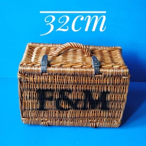 32cm Fortnum and Mason willow picnic basket. The basket is sitting upright, with the Fortnum and Mason logo facing front. The basket has an integrated willow handle, plus a hinged lid that has two leather straps to hold the lid securely in-place