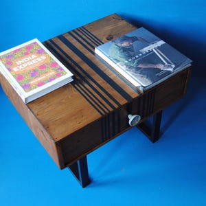 May include: A square wooden side table with a natural brown finish and black vertical stripes across the top and front drawer. The table features a single drawer with a small white knob and is supported by black metal legs. Two books rest on top.