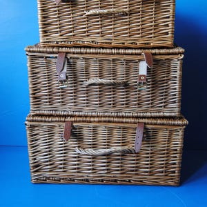selection of different sized Fortnum and Mason rectangular hamper baskets. Baskets are arranged in order of size, with the largest at the bottom. The baskets are made of brown willow. They have carry handles at the front. Also visible are the leather closing straps