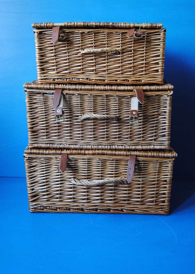 selection of different sized Fortnum and Mason rectangular hamper baskets. Baskets are arranged in order of size, with the largest at the bottom. The baskets are made of brown willow. They have carry handles at the front. Also visible are the leather closing straps