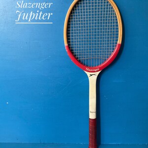 A vintage wooden Slazenger Jupiter tennis racket. The racket is predominantly in cream and burgundy. The Slazenger logo is visible. The racket is shown against a blue background