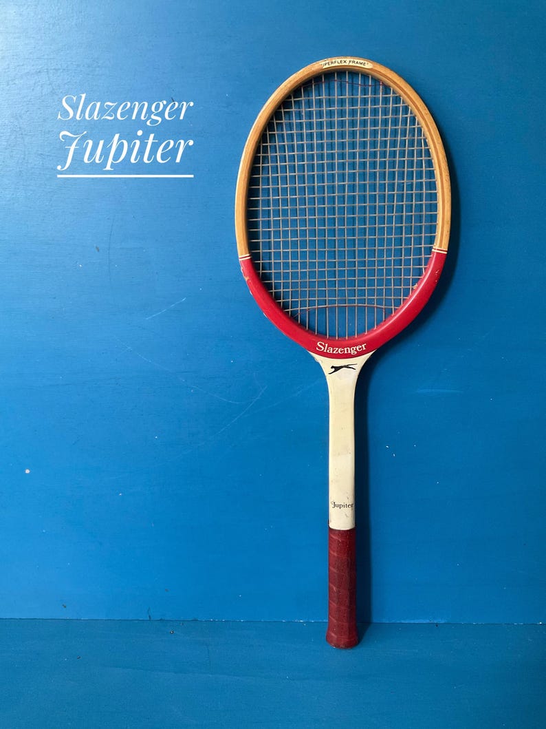 A vintage wooden Slazenger Jupiter tennis racket. The racket is predominantly in cream and burgundy. The Slazenger logo is visible. The racket is shown against a blue background